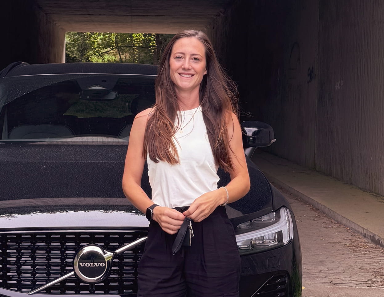 Kim Irmgartz steht vor einem Volvo‑Auto. Sie posiert vor dem Fahrzeug, das im Vordergrund gut sichtbar ist, während der Hintergrund eine klare Außenaufnahme zeigt.