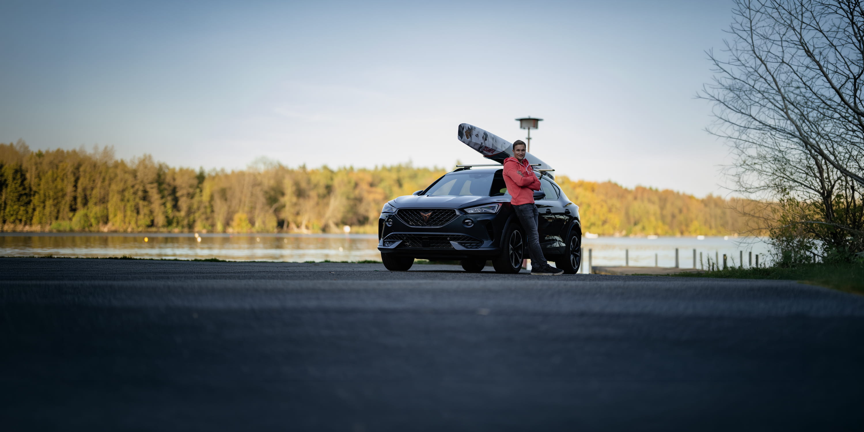 Ein Kanufahrer steht vor seinem Auto, auf dessen Dach ein Kanu sicher befestigt ist. Er posiert vor dem Fahrzeug, während das Kanu gut sichtbar auf dem Dachträger liegt.