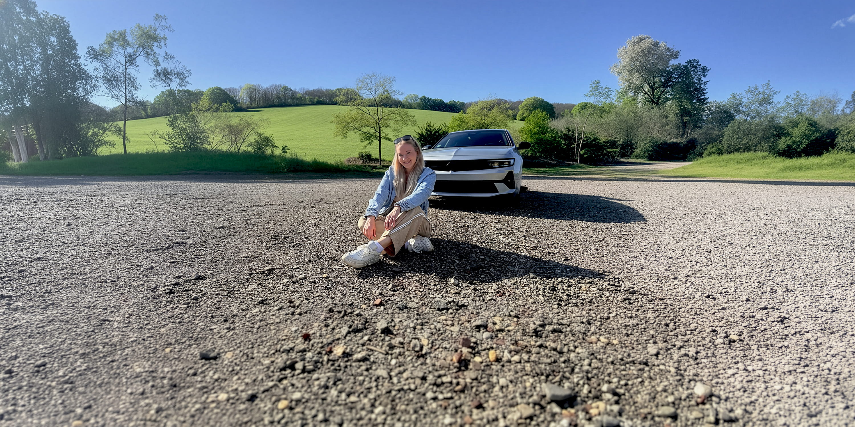 Aileen Tina Hufschmidt sitzt vor einem Auto und posiert für ein Foto. Sie ist klar im Vordergrund zu sehen, während das Fahrzeug und die Umgebung den Hintergrund bilden.