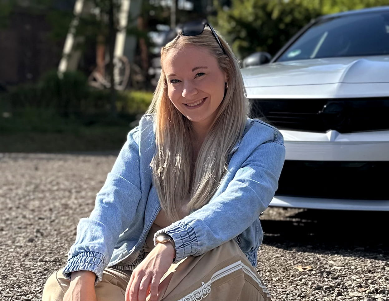 Aileen Tina Hufschmidt sitzt vor einem Auto und posiert für ein Foto. Sie ist klar im Vordergrund zu sehen, während das Fahrzeug und die Umgebung den Hintergrund bilden.